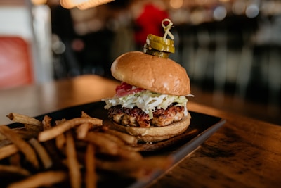 A gourmet burger with a juicy patty topped with lettuce, pickles, and onions sits on a dark plate. The burger is accompanied by a side of golden-brown French fries. The background is softly blurred, suggesting a cozy dining atmosphere.