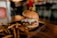 A gourmet burger with a juicy patty topped with lettuce, pickles, and onions sits on a dark plate. The burger is accompanied by a side of golden-brown French fries. The background is softly blurred, suggesting a cozy dining atmosphere.