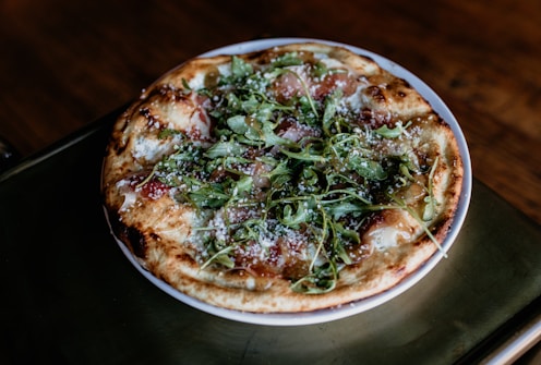 A freshly baked pizza topped with greens, possibly arugula, and sprinkled with cheese rests on a round white plate. The crust appears golden brown, with slight charring, and the toppings are distributed evenly across the surface.