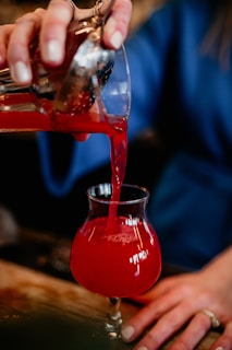A close-up of a hand pouring a craft spirit into a glass with a California sunset in the background.