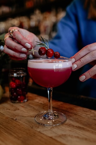 A person is delicately placing a garnish of cranberries and rosemary on a pink cocktail in a coupe glass. The scene appears to be set in a bar with a wooden countertop, against a blurred background filled with various bar elements.