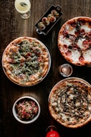 A variety of delicious foods including three different pizzas, a bowl of salad, deviled eggs on a small tray, a cocktail, and a candle are artfully arranged on a dark wooden table.