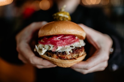 A person holding a hamburger with toppings including sliced red onions, cabbage, and a beef patty, while a pickle is skewered on top. The background is softly blurred with warm lighting, focusing attention on the burger.