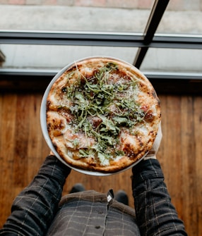 A person is holding a pizza topped with arugula and some grated cheese, standing on a wooden floor. The pizza appears freshly baked with a crispy crust, and the person is wearing a checkered shirt.