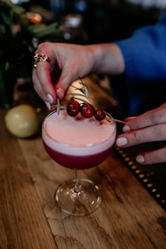 A person garnishes a cocktail with a skewer of red berries and a sprig of rosemary. The drink is served in a coupe glass with frothy foam on top, set on a wooden table.