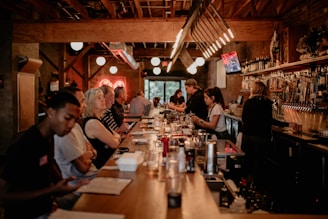 A cozy bar setting with people seated along a wooden counter. The dim lighting creates a warm and intimate atmosphere, enhanced by hanging lights and a neon sign in the background. Bottles and taps line the bar, and bartenders are busy attending to customers.