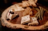 A wooden platter with an assortment of gourmet foods. There are several varieties of cheese, with one piece topped with some blackberries and drizzled with a dark syrup. Additionally, there are green beans laid out alongside honeycomb and toasted bread slices.