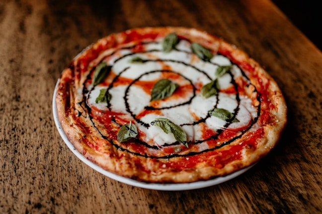 A freshly baked pizza sits on a wooden table. The pizza has a thin crust, topped with melted mozzarella cheese, tomato sauce, fresh basil leaves, and drizzled with a balsamic glaze creating a spiral pattern.