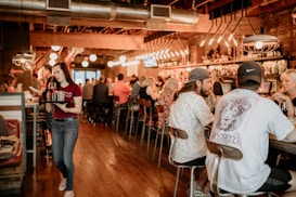 A bustling bar or restaurant setting with people sitting at bar stools engaged in conversation. A waitress wearing a red shirt and jeans carries a tray with drinks. The atmosphere is warm with ambient lighting, exposed ductwork, and wooden flooring.