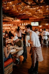 A cozy, warmly lit restaurant interior with a wooden ceiling adorned with string lights. Patrons are engaging in conversation while enjoying their meals. A server, casually dressed, is attentively interacting with a seated couple. The space conveys a lively and inviting atmosphere.