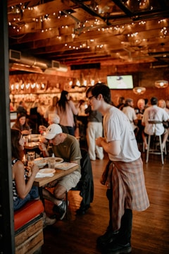 A cozy, warmly lit restaurant interior with a wooden ceiling adorned with string lights. Patrons are engaging in conversation while enjoying their meals. A server, casually dressed, is attentively interacting with a seated couple. The space conveys a lively and inviting atmosphere.
