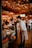 A cozy, warmly lit restaurant interior with a wooden ceiling adorned with string lights. Patrons are engaging in conversation while enjoying their meals. A server, casually dressed, is attentively interacting with a seated couple. The space conveys a lively and inviting atmosphere.