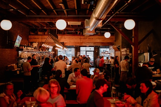A bustling bar or restaurant with patrons seated at tables and standing at the bar. The space features warm, dim lighting with exposed brick walls and a wooden ceiling. People are engaged in conversation, enjoying drinks, and watching a television screen displaying a sporting event. The atmosphere is lively and sociable.