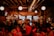 A bustling bar or restaurant with patrons seated at tables and standing at the bar. The space features warm, dim lighting with exposed brick walls and a wooden ceiling. People are engaged in conversation, enjoying drinks, and watching a television screen displaying a sporting event. The atmosphere is lively and sociable.