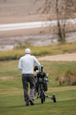 A person wearing a white cap and light-colored sweater is walking on a golf course, pushing a golf cart. The background includes a grassy area and a sand bunker, with a hazy, autumnal scene in the distance.