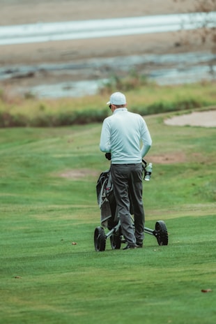 A person wearing a white long-sleeved shirt and dark pants is walking on a well-maintained grassy field with a wheeled golf bag. The background shows a body of water with a sandy shore and some foliage.