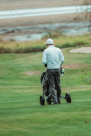A person wearing a white long-sleeved shirt and dark pants is walking on a well-maintained grassy field with a wheeled golf bag. The background shows a body of water with a sandy shore and some foliage.
