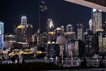 A city skyline at night featuring numerous high-rise buildings illuminated by various lights creates a vibrant and dynamic atmosphere. Foreground elements include circular street lamps and people casually gathering or walking along a railing.