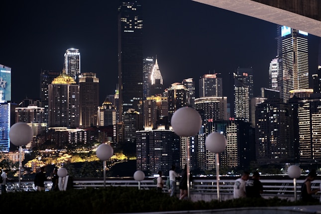 A city skyline at night featuring numerous high-rise buildings illuminated by various lights creates a vibrant and dynamic atmosphere. Foreground elements include circular street lamps and people casually gathering or walking along a railing.