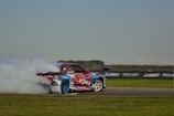 A racing car is drifting on a track, emitting a large cloud of white smoke from its tires. The car is decorated with vibrant decals in red, blue, and white, and is positioned against a backdrop of a grassy field and a clear sky.