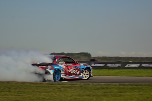 A racing car is drifting on a track, emitting a large cloud of white smoke from its tires. The car is decorated with vibrant decals in red, blue, and white, and is positioned against a backdrop of a grassy field and a clear sky.