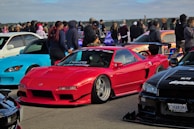 A vibrant car meet featuring several modified sports cars. A red sports car takes center focus, accompanied by a blue convertible on the left and a sleek black car on the right. Several people gather around the vehicles, some dressed in casual attire, suggesting a social gathering.