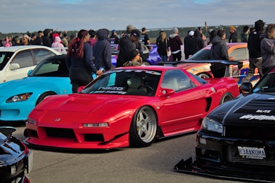 A vibrant car meet featuring several modified sports cars. A red sports car takes center focus, accompanied by a blue convertible on the left and a sleek black car on the right. Several people gather around the vehicles, some dressed in casual attire, suggesting a social gathering.