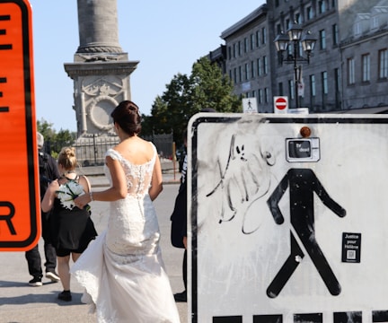 A woman in a white wedding dress is walking down a street, accompanied by another person holding a bouquet. There is a large, decorated stone column with historical carvings in the background. To the right, a street sign features an altered pedestrian symbol with the phrase 'tv dead'. Buildings line the street, and a lamp post is visible.