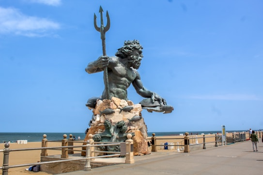 A large statue of a powerful figure wielding a trident, sitting atop a rocky pedestal surrounded by sea creatures, positioned along a beachfront promenade. The background features a sandy beach with a few people and a calm, blue ocean under a clear sky.