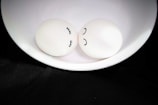 Two eggs with drawn faces rest in a white bowl. One egg has eyelashes and a serene expression, while the other has a simple curved line as a smile. They are presented on a black fabric background, creating a contrast between the softness of the eggs and the dark surroundings.