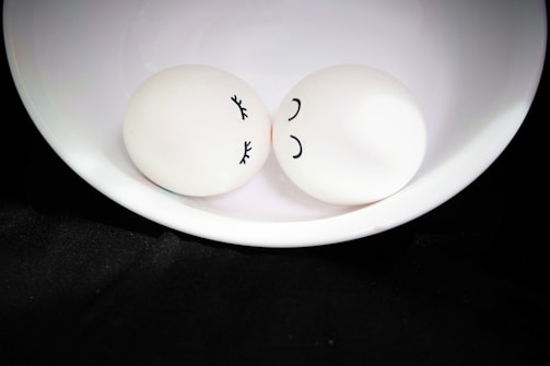 Two eggs with drawn faces rest in a white bowl. One egg has eyelashes and a serene expression, while the other has a simple curved line as a smile. They are presented on a black fabric background, creating a contrast between the softness of the eggs and the dark surroundings.