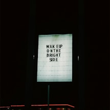 A lighted signboard with the text 'WAKE UP ON THE BRIGHT SIDE' is prominently displayed against a dark background. The sign is illuminated and the surrounding area is mostly dark, with a hint of red neon light at the bottom edge.
