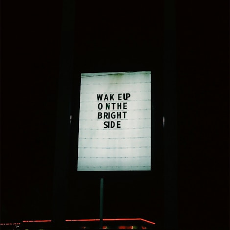 A lighted signboard with the text 'WAKE UP ON THE BRIGHT SIDE' is prominently displayed against a dark background. The sign is illuminated and the surrounding area is mostly dark, with a hint of red neon light at the bottom edge.