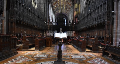 A grand, ornate cathedral interior with high vaulted ceilings. The aisle is flanked by intricately carved wooden choir stalls, and an open book rests on a stand in the center. The flooring features detailed geometric patterns, and the ambient light creates a serene atmosphere.