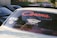 Vintage car with stickers on the rear windshield, prominently featuring the word 'Corona' in bold red font with decorative elements. Other stickers include 'WildRetro' and a circular design. The car is surrounded by other vehicles and greenery can be seen in the background.