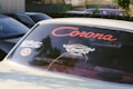 Vintage car with stickers on the rear windshield, prominently featuring the word 'Corona' in bold red font with decorative elements. Other stickers include 'WildRetro' and a circular design. The car is surrounded by other vehicles and greenery can be seen in the background.