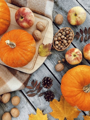 An arrangement of autumn-themed objects including pumpkins, apples, walnuts, acorns, and pinecones on a rustic wooden surface. A cutting board and a plaid cloth add a cozy touch, while scattered dried leaves enhance the fall atmosphere.