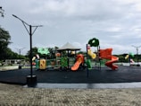 A vibrant playground featuring colorful slides, climbing structures, and swings, set in an open outdoor park area. The ground is covered with black safety padding, and there are cobblestone pathways surrounding the play area. The sky is overcast, and there are trees in the background, providing a natural setting.