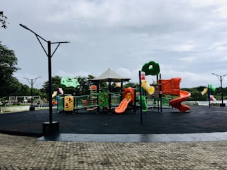 A vibrant playground featuring colorful slides, climbing structures, and swings, set in an open outdoor park area. The ground is covered with black safety padding, and there are cobblestone pathways surrounding the play area. The sky is overcast, and there are trees in the background, providing a natural setting.