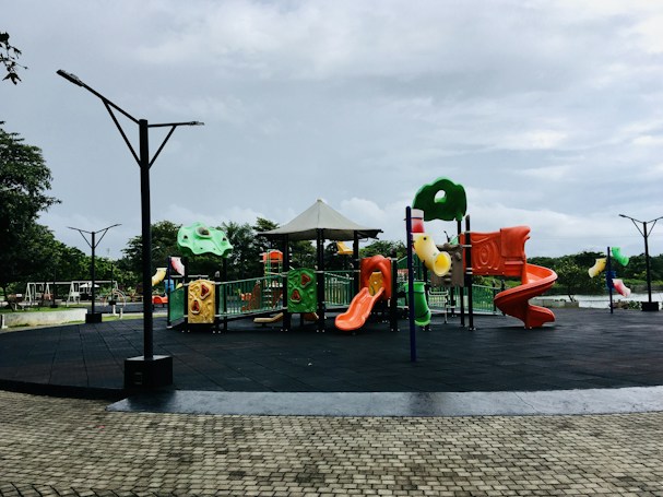 A vibrant playground featuring colorful slides, climbing structures, and swings, set in an open outdoor park area. The ground is covered with black safety padding, and there are cobblestone pathways surrounding the play area. The sky is overcast, and there are trees in the background, providing a natural setting.