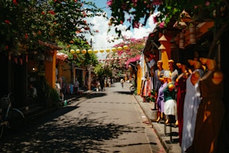 A vibrant street scene lined with shops on both sides, featuring colorful mannequins displaying dresses on the right side and lush flowering trees overhead. Lanterns hang across the narrow street, casting shadows on the ground, while people are seen casually walking and biking in the background. The atmosphere suggests a lively, bustling market area with a traditional charm.