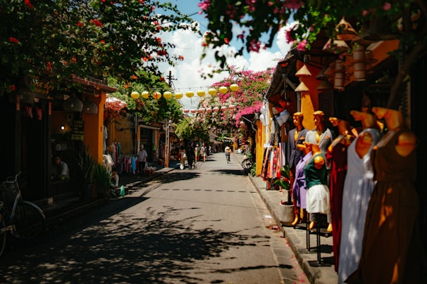 A vibrant street scene lined with shops on both sides, featuring colorful mannequins displaying dresses on the right side and lush flowering trees overhead. Lanterns hang across the narrow street, casting shadows on the ground, while people are seen casually walking and biking in the background. The atmosphere suggests a lively, bustling market area with a traditional charm.