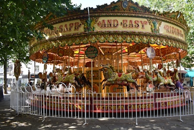A vintage carousel with ornate decorations and colorful horses, surrounded by a white fence, set under a canopy of leafy trees. The carousel features intricate patterns and bright lights, creating a nostalgic and whimsical atmosphere.