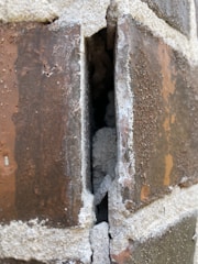 A close-up view of a brick wall with a visible crack or gap between the bricks, revealing some of the material inside. The surface of the bricks appears weathered and is covered with a white, powdery substance.