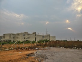 A large institutional building with multiple stories is set against a cloudy sky. The building is surrounded by an open area with dried grass and sparse greenery, enclosed by a simple wire fence. Electric poles and wires are visible, adding to the industrial feel.