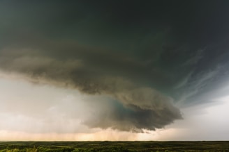 A massive storm cloud dominates the sky, swirling with dense, dark formations over an open, grassy plain. The cloud appears heavy with rain, and the atmosphere is charged with the impending sense of a storm.