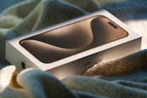 A boxed smartphone rests on a soft, textured fabric under warm, natural lighting. The packaging features a sleek and minimalistic design with a sand dune-like pattern on the top.