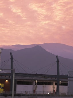 A modern building with large structural beams stands in the foreground, silhouetted against a backdrop of mountains. Above, the sky is filled with soft, pastel-colored clouds, creating a serene atmosphere.
