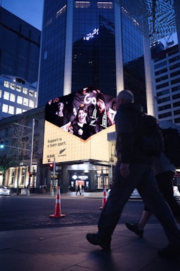 A city street scene featuring a large, illuminated billboard on the side of a building, displaying an advertisement with cheering sports fans and the text 'Support is a team sport.' People are walking past in the foreground, while traffic cones are placed on the sidewalk. The scene suggests an urban environment in the evening.