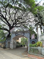 An entrance archway to Dhaka Medical College is surrounded by lush greenery and covered with several posters. There are trees overhead with sprawling branches, creating a canopy. Two rickshaws are parked inside the gate, indicative of a busy, urban setting.